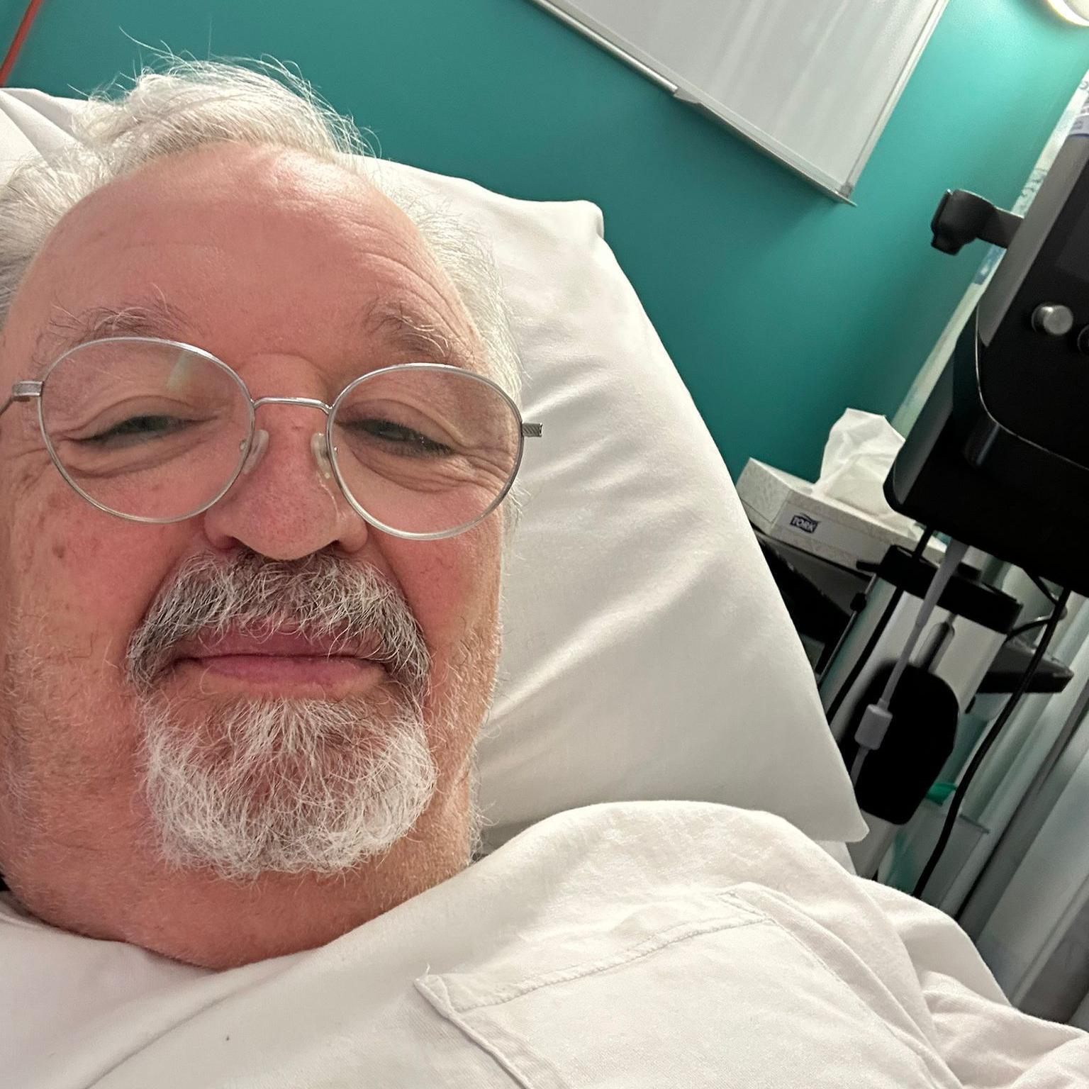 Nigel McCrery taking a selfie while lying his head on a pillow. He has a white beard and is wearing a white t-shirt. He was grey-rimmed glasses on.