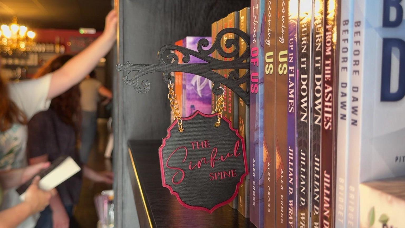 A bookshelf with a small black sign stuck among the books, saying The Sinful Spine in cursive red script. In the background, people can be seen reaching for the books, but they are out of focus.