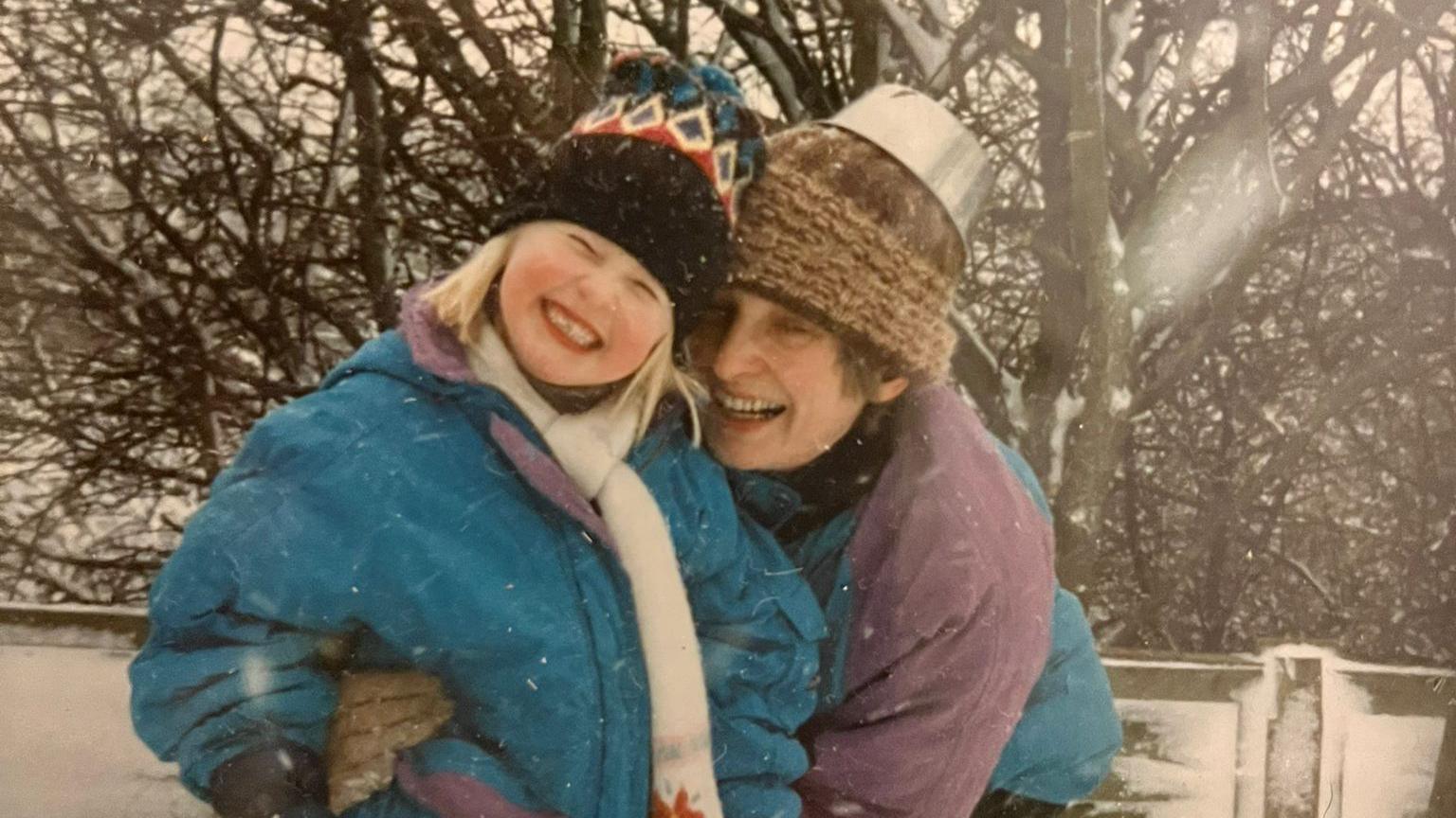 A child in a blue coat wearing a white scarf and black chequered hat is cuddled by a woman in a purple and blue coat with a brown hat. They are smiling and it is snowing.