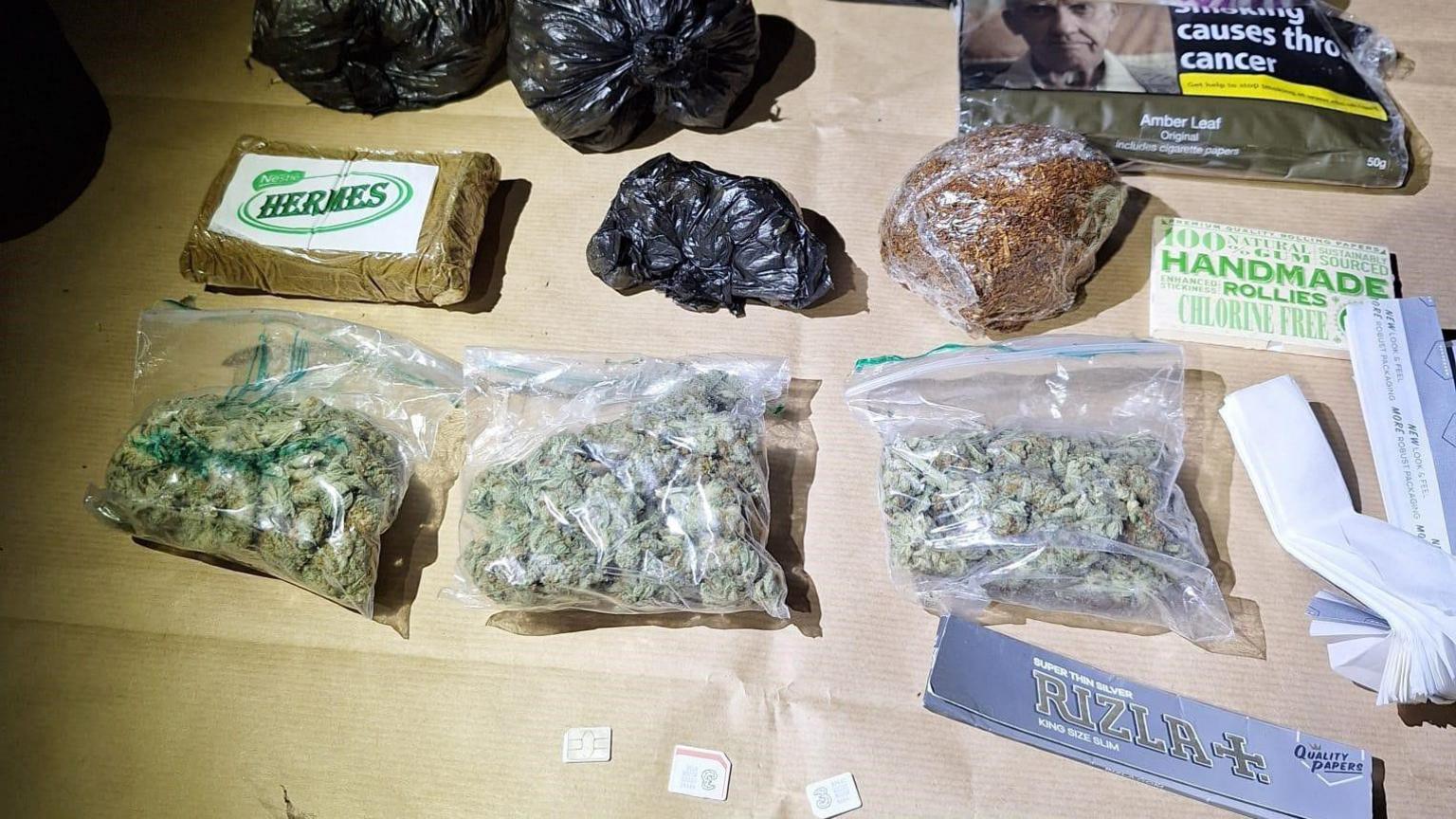 Clear plastic snap bags filled with cannabis are lined up on a table. Black bin bag-style wraps are also seen displayed, alongside cigarette rolling papers, a pouch of tobacco and three phone SIM cards. 