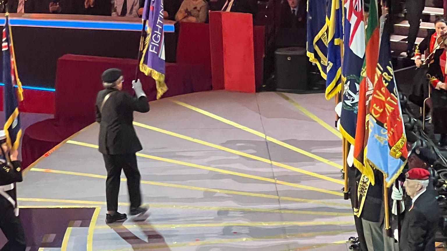 A woman wearing a suit and beret, holding a flag, while walking across a stage to a row of veterans, each holding their regimental flag