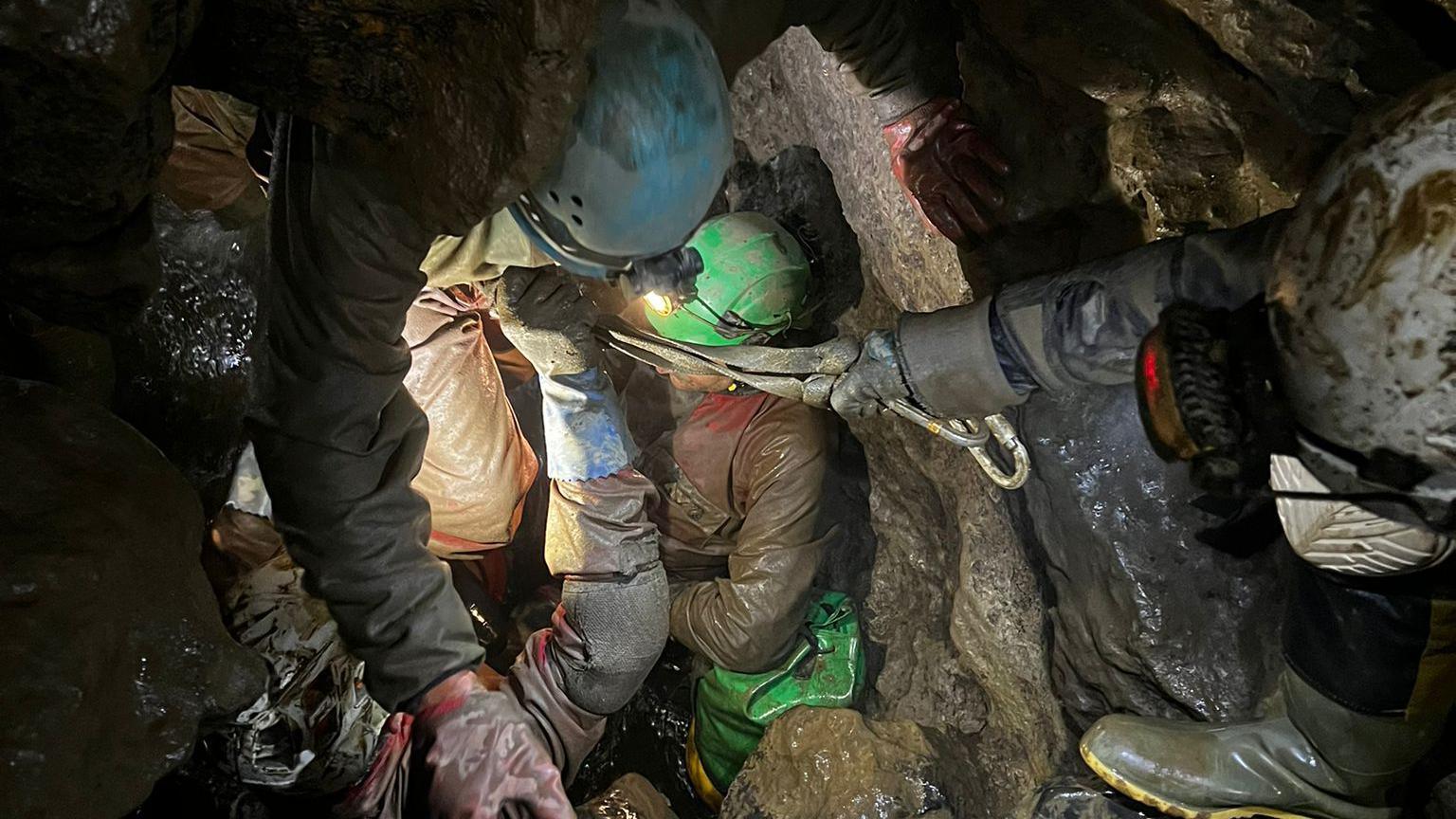 A group of people navigate through a narrow, rocky passage within a cave, dressed in appropriate gear including helmets with headlamps, gloves, and protective clothing. One individual is assisting another by holding onto a rope or harness. The cave walls appear wet and textured, and the scene is primarily lit by the headlamps, which cast light on the immediate surroundings.
