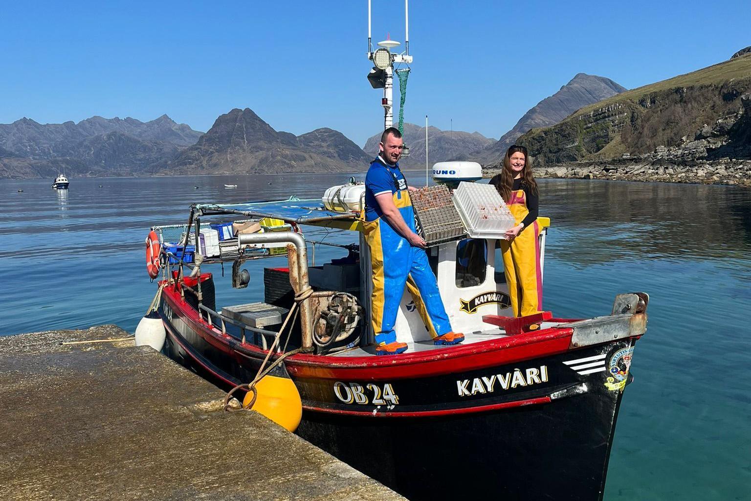 Brooke Lamond stands on a small creel fishing boat with James Robertson. She wears a long-sleeve black top with yellow and pink fishing overalls on top. James wears a short-sleeve blue top with blue and yellow fishing overalls. It is a sunny day and you can see the still water around them and hills in the distance.