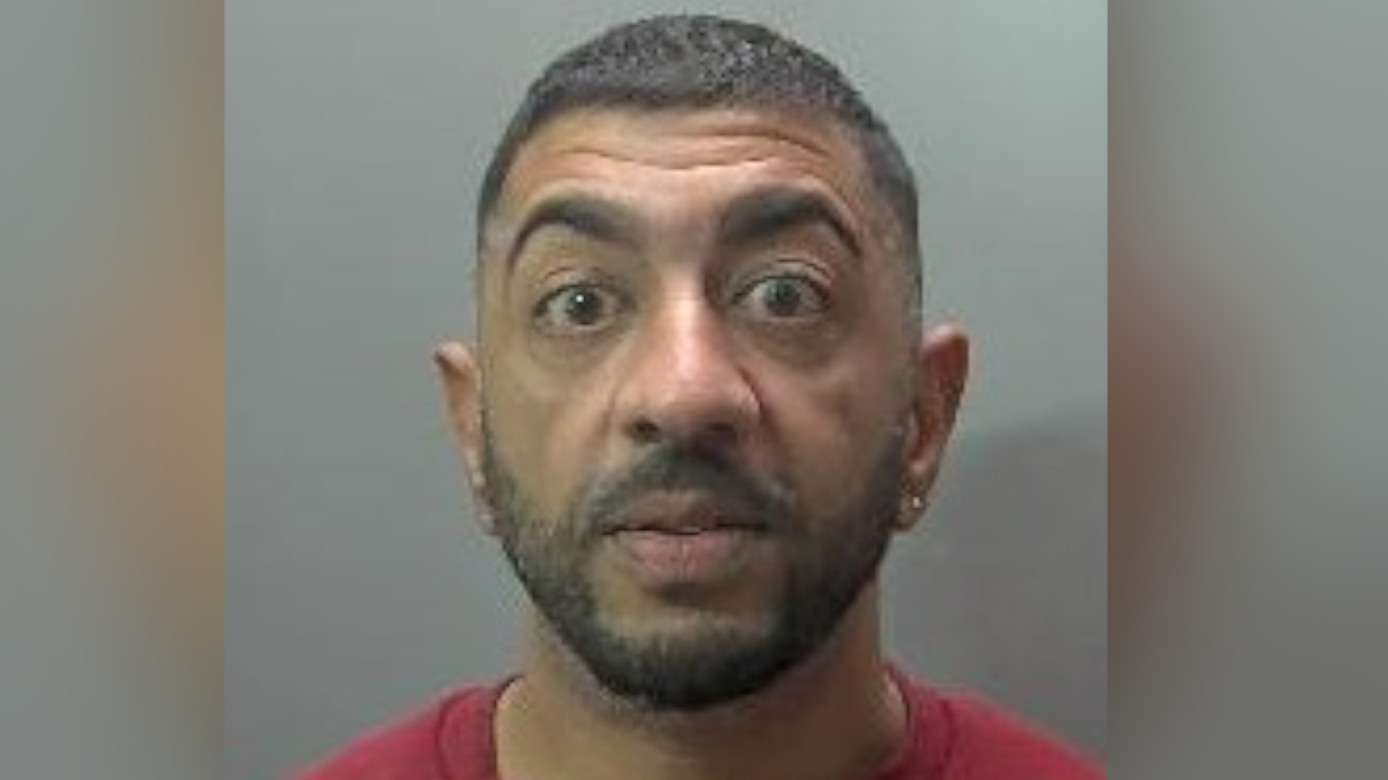 Custody image of Ali Amjad against a grey background. He has an earing in his right ear and dark short hair and a dark beard. He is wearing a red top. His eyebrows are raised so he appears shocked.