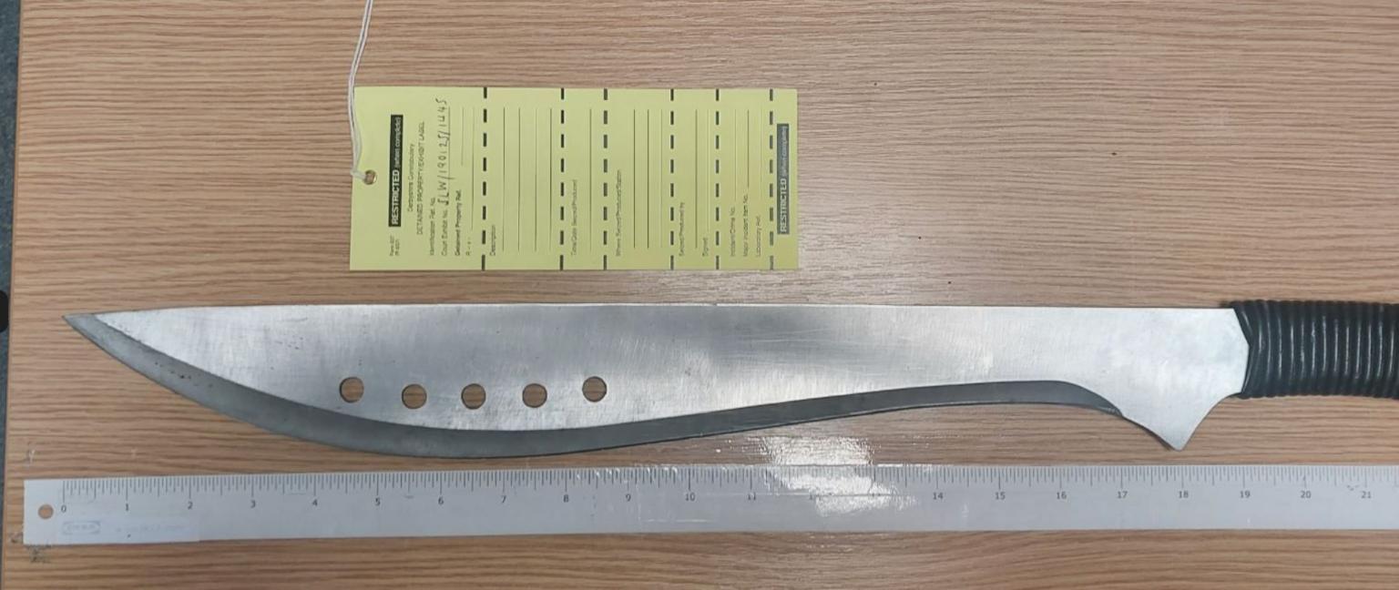 A silver machete with black handle on a wooden surface with a ruler next to it