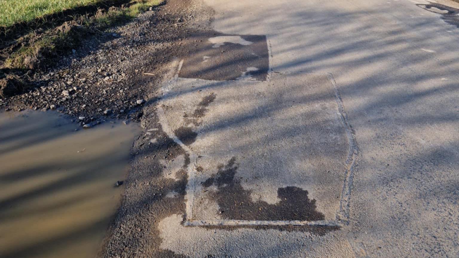 A side view of a road showing a large watery puddle to the left and a patched-up pothole in the road to the right