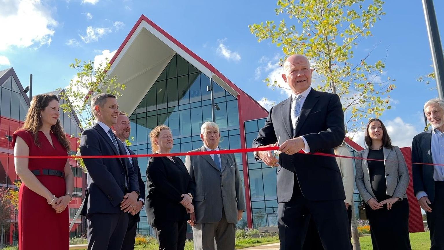 Lord Hague is wearing a dark suit and light blue tie and is holding scissors in one hand, and a red ribbon in the other.  Behind him is a brand new building with glass walls and a red roof.  A group of smartly dressed people are gathered behind him.