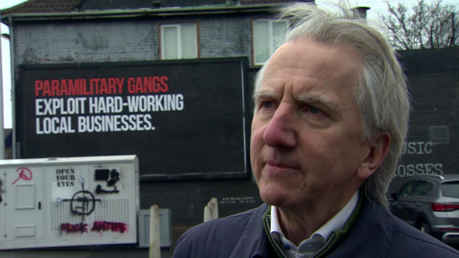 Máirtín Ó Muilleoir with a paramilitary sign in the background. He has white hair, wearing a blue jacket.