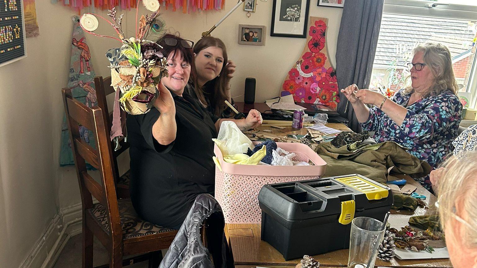 A group of women participating in a craft workshop, sitting round a table with lengths of cloth and loose craft supplies on it 