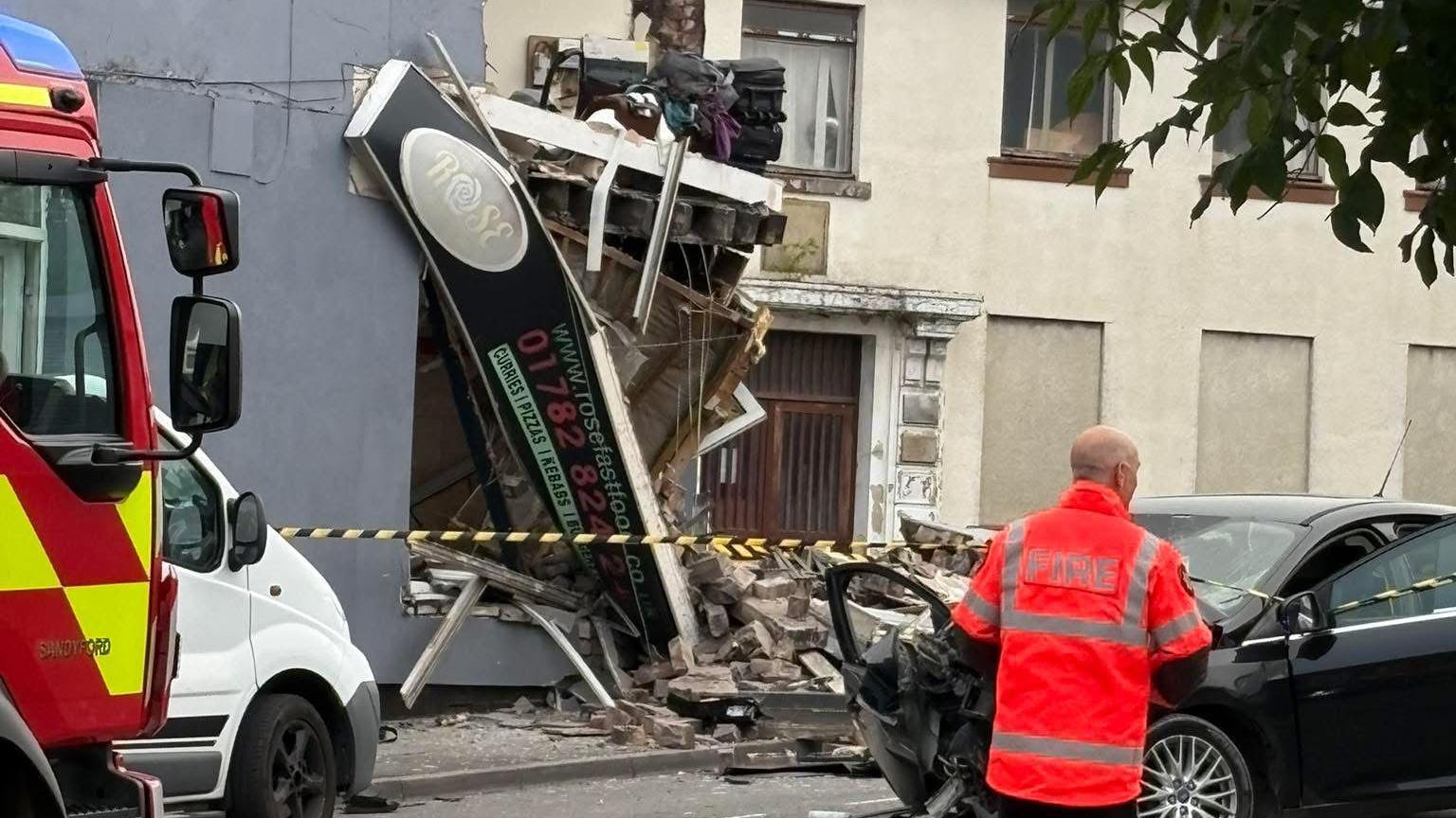A partially collapsed shop, with rubble on the street and the shop sign hanging down. Police tape is around it. A man in a bright orange hi-vis jacket stands in front of a black car, which has damage to its front and both doors open. The red and yellow chequered pattern at the front of a fire and rescue service vehicle can be seen on the left.