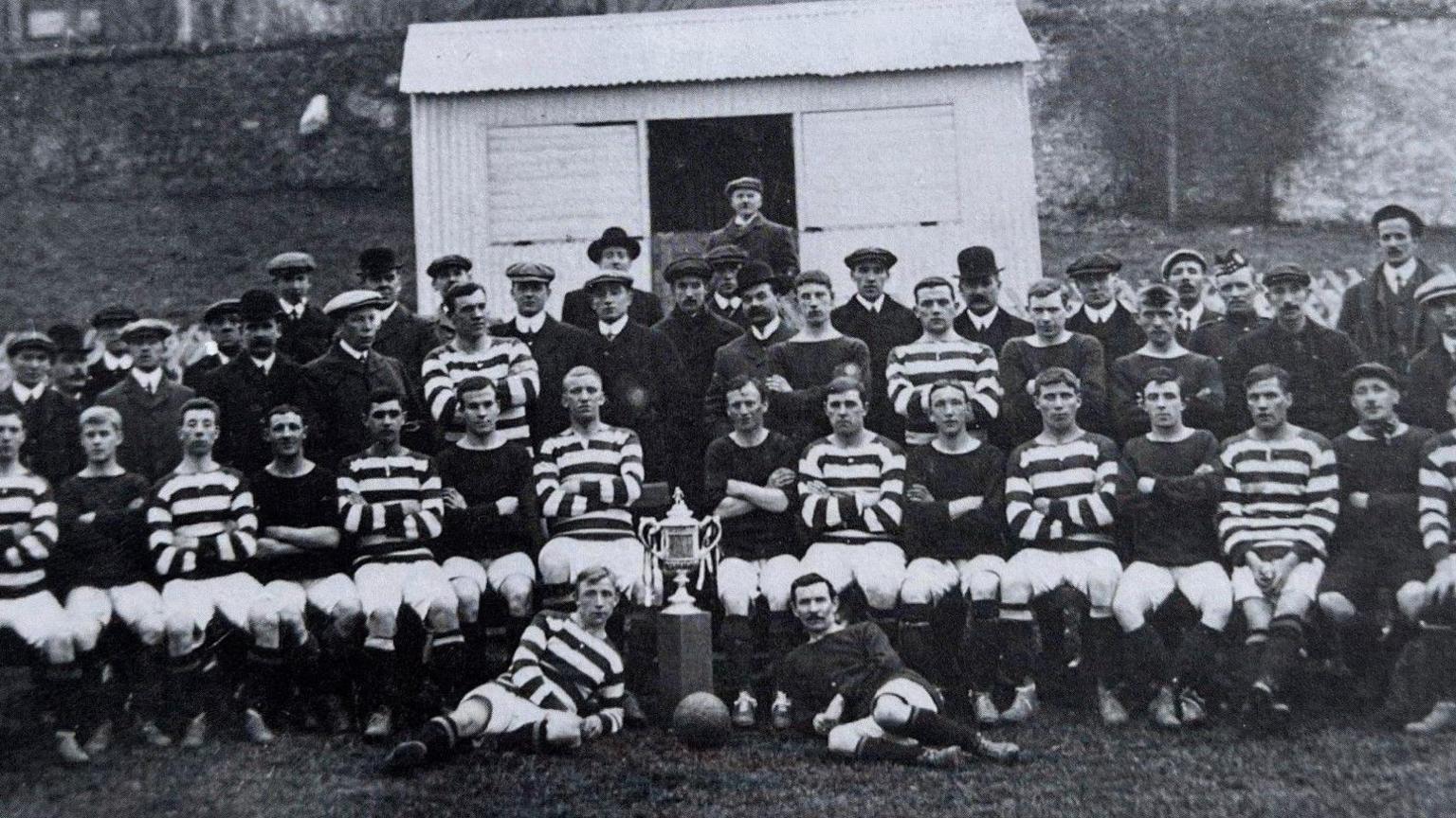 A large group of football players, along with coaches, line up in a black-and-white photograph from 1908. A large trophy takes centre stage