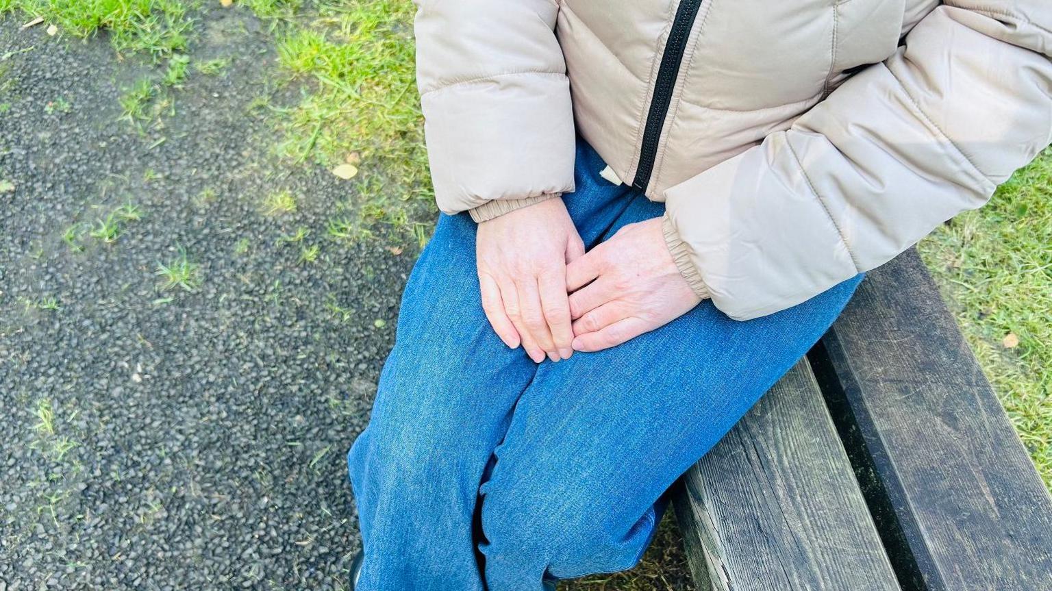 A woman's hands clasped together. She wears jeans and a beige puffer coat