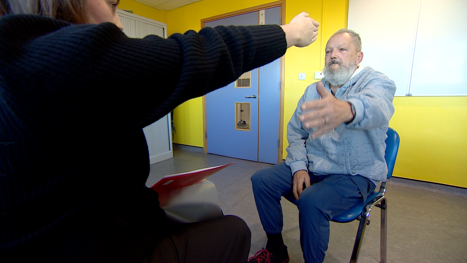 Michael Koers and the doctor sit opposite each other during a rehabilitation session. Michael and the doctor have their arms in the air. 