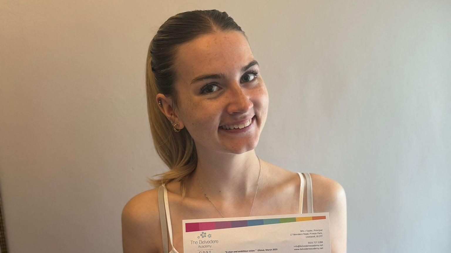 Lily-Rose Williamson, a young woman with long blonde hair tied back in a ponytail, smiles for the camera in front of a plain white wall. She is holding a sheet of paper with her A-level results printed on it, the top of which is visible in the image.