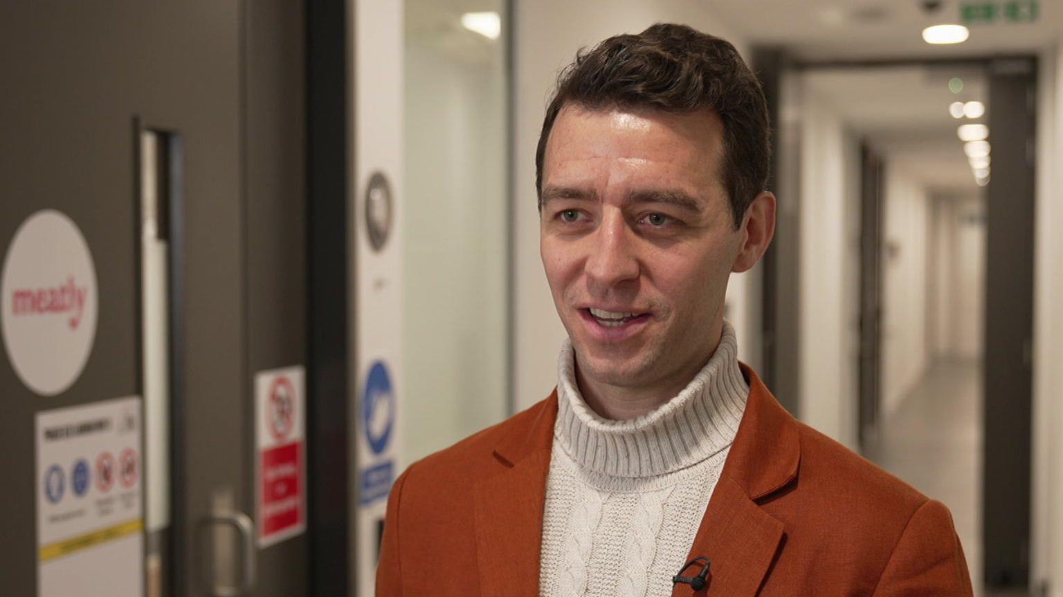 Owen Ensor stands in a corridor giving a TV interview. He is 36 years old, has brown, wavy hair and wears a white roll-neck jumper and dark orange jacket.