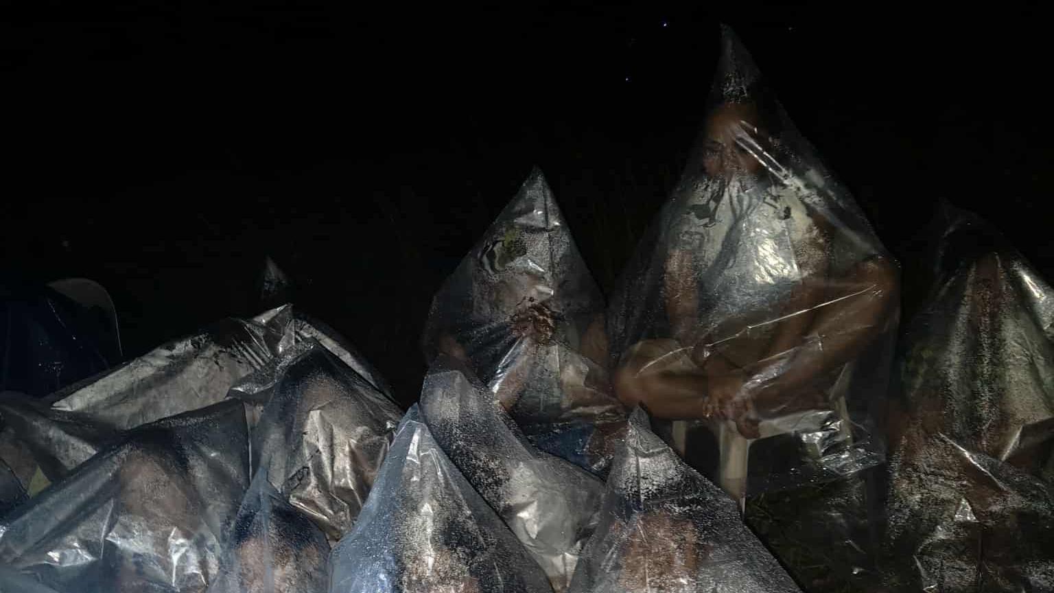 People covered in plastic bags, which have fogged up with their breath, sitting on stools in the dark