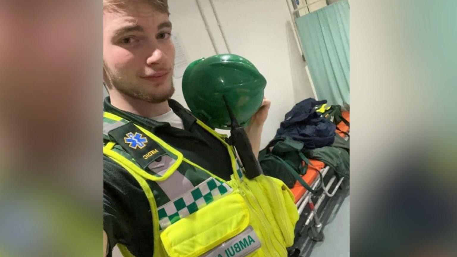 Jamie Kadolski poses in a selfie wearing an high-visibility vest with "ambulance" written on it. Underneath the vest is a green polo shirt and he is holding a green helmet. He is has a radio attached to the vest. In the background is a stretcher with orange mattress with bags on top. 