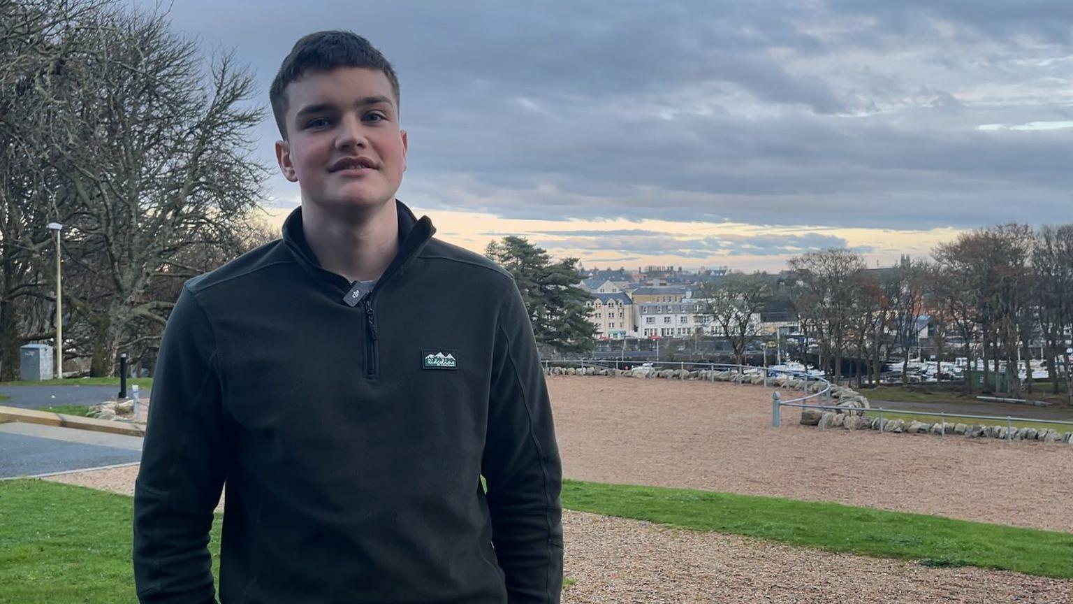 Seumas Mac a' Ghobhainn, air a bheil Type 1, na sheasamh le gàire air aodann agus baile Steòrnabhaigh air a chùl 