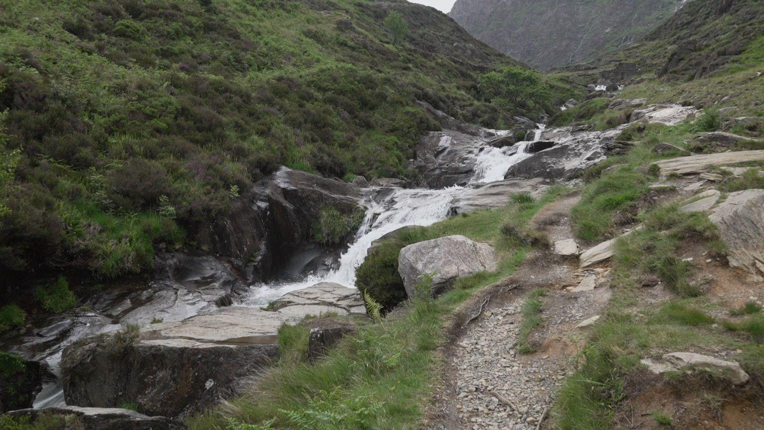 A stream cascades down a series of rocks alongside a walking path. The bank slopes upwards on the left hand side.
