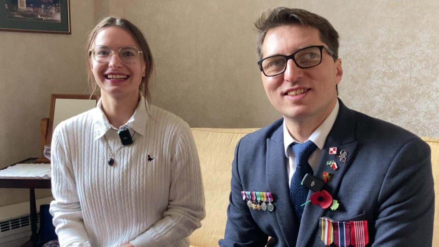 Agata Koralewska and Andrzej Michalski sit on a yellow sofa. Andrzej is wearing a blazer with a poppy and military medals on. Agata is wearing a white jumper.