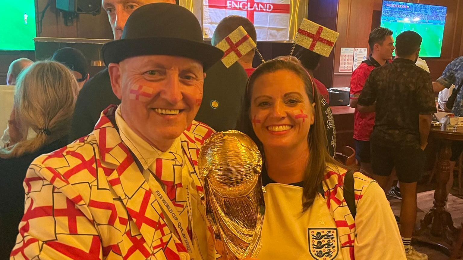 A man wearing a St George suit covered in red and white England flags and a black bowler hat smiles for a photo with a woman wearing an England shirt. The pair are holding up a replica golden World Cup.