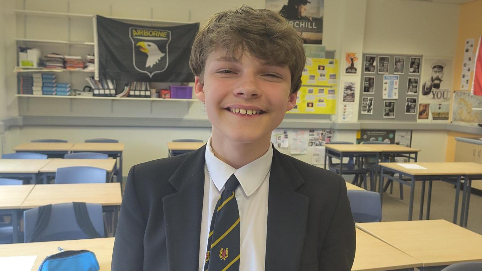 Ethan is standin in a classroom smiling into the camera. He is wearing a school uniform