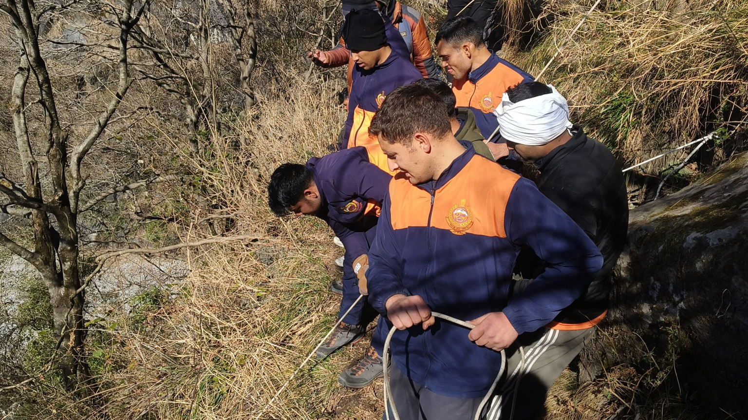 Rescue workers wearing blue and orange fleeces hold ropes as they work to tether the stretcher to the mountain