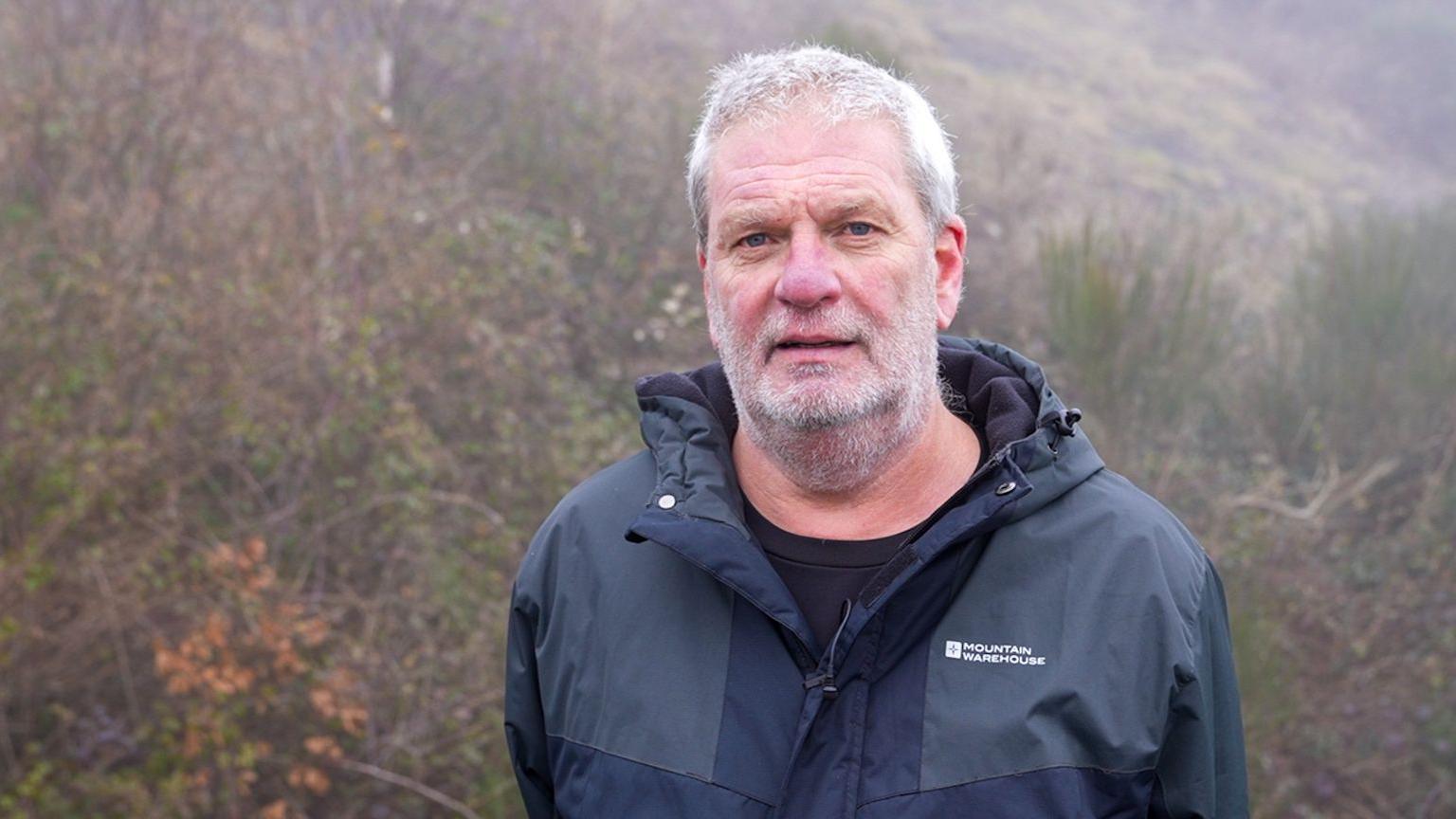 Stuart Adams looking at the camera. He has grey hair and a grey beard (stubble) and moustache. He is wearing a blue waterproof coat and is standing in front of trees. 