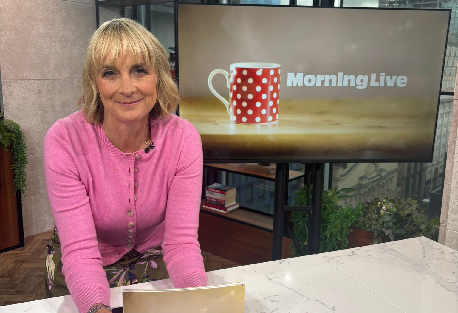Louise Minchin leaning on the breakfast bar in the Morning Live studio holding cue cards.