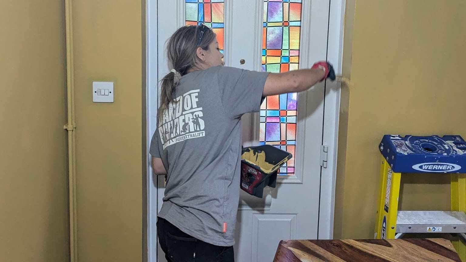 A tradeswoman wearing a grey t-shirt with "Band of Builders" printed on the back. She is painting yellow onto a wall by a front door.