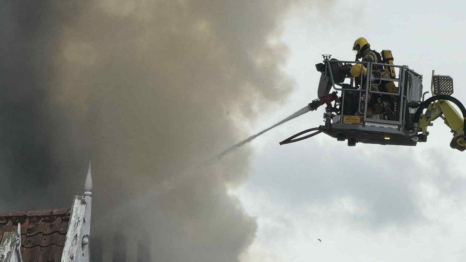 Using an aerial ladder, fire crews tackle a fire. They are using a hose and there is a large plume of smoke.