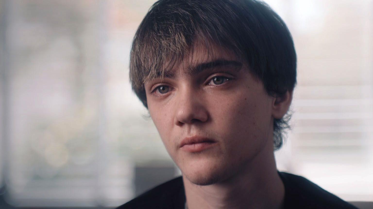 
A close-up of a young man indoors, with short hair, looking towards the camera.
