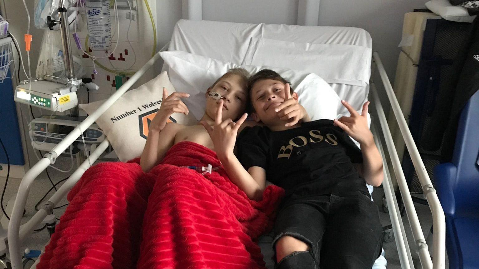 Ben lying in his hospital bed, with a red blanket covering him. His brother Jack is lying next to him while wearing black Hugo Boss T-shirt.