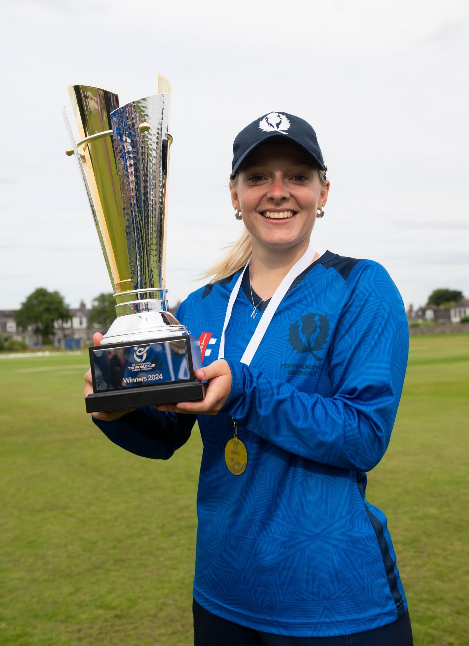 Niamh Muir, with blonde hair, is in a Scotland top and wearing a blue cap with a thistle on it. She is holding up a large trophy while wearing a medal around her neck