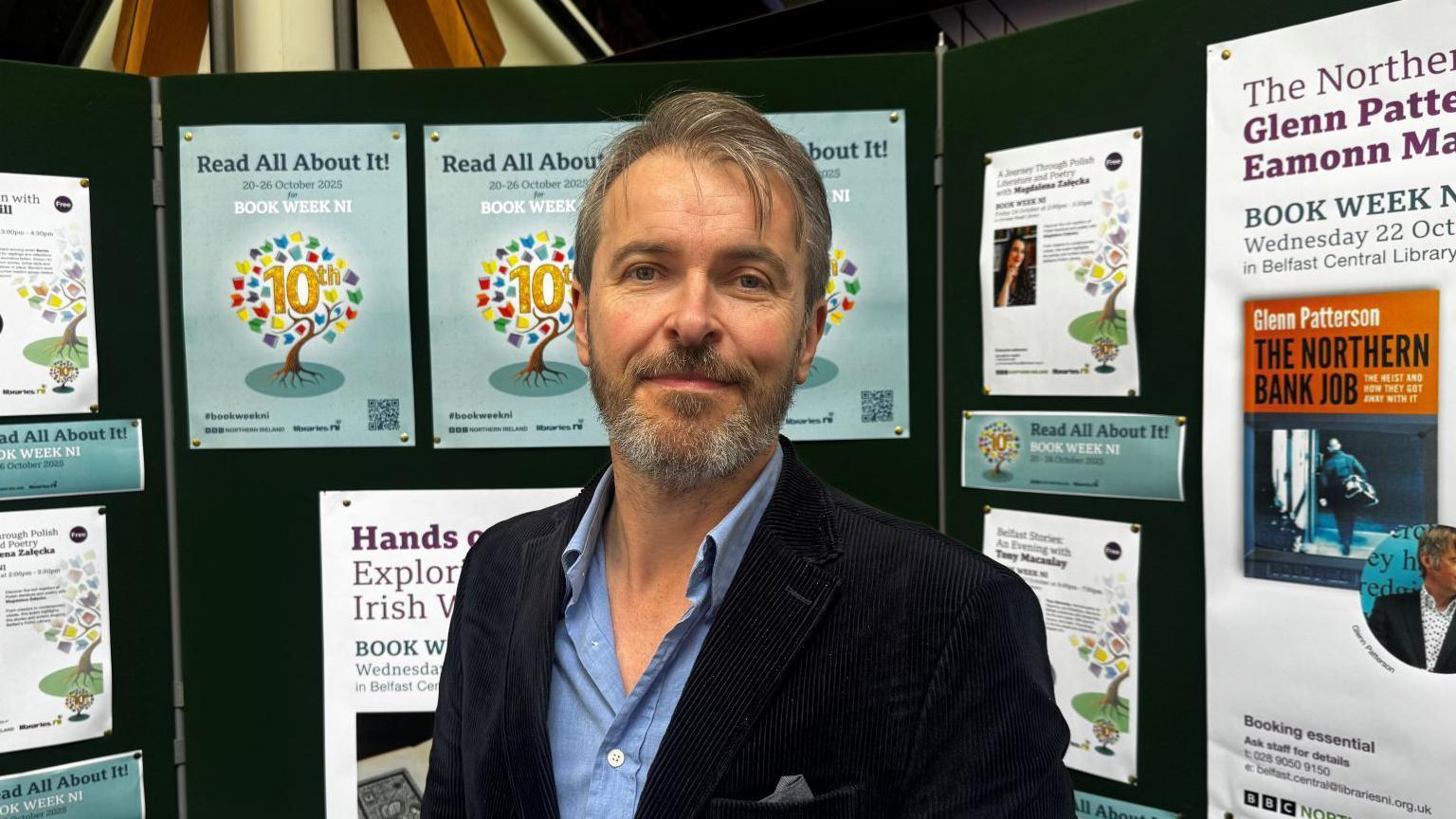 Garett Carr wearing a blue shirt and navy blazer is smiling at the camera. There are posters for book week NI in the background. 