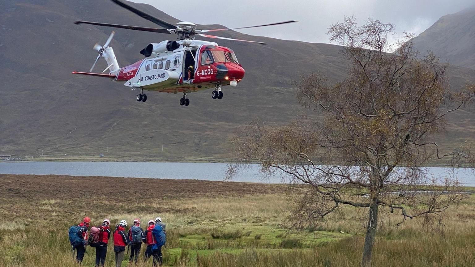 A red and white helicopter landing on a green strip of grass with mountains and a loch behind.