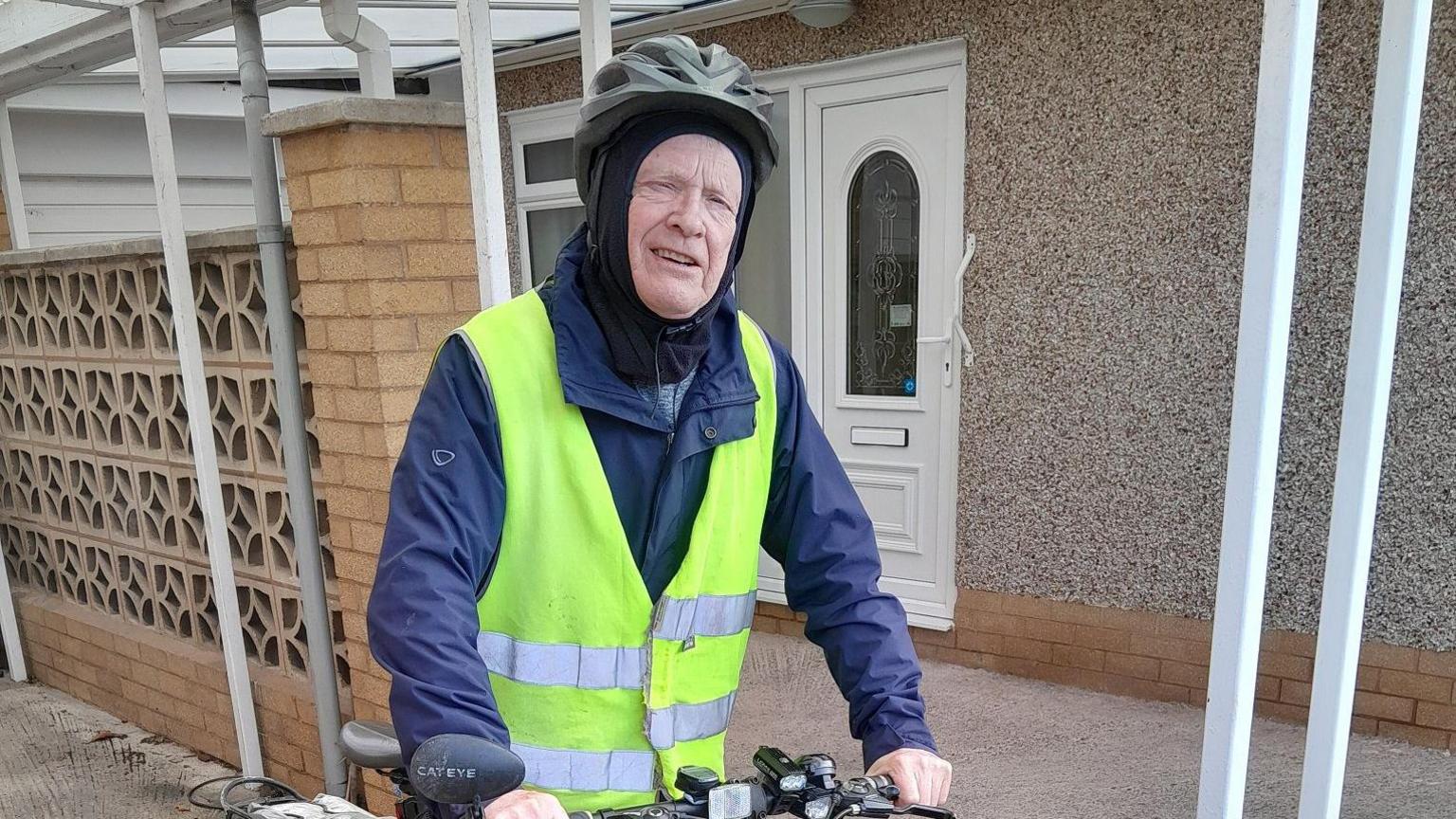 Tudur Davies, 78 ar ei feic yn barod i fynd i dynnu lluniau.  Mae'n gwisgo cot las gyda siaced felyn llachar drosti a helmet ddu am ei ben.