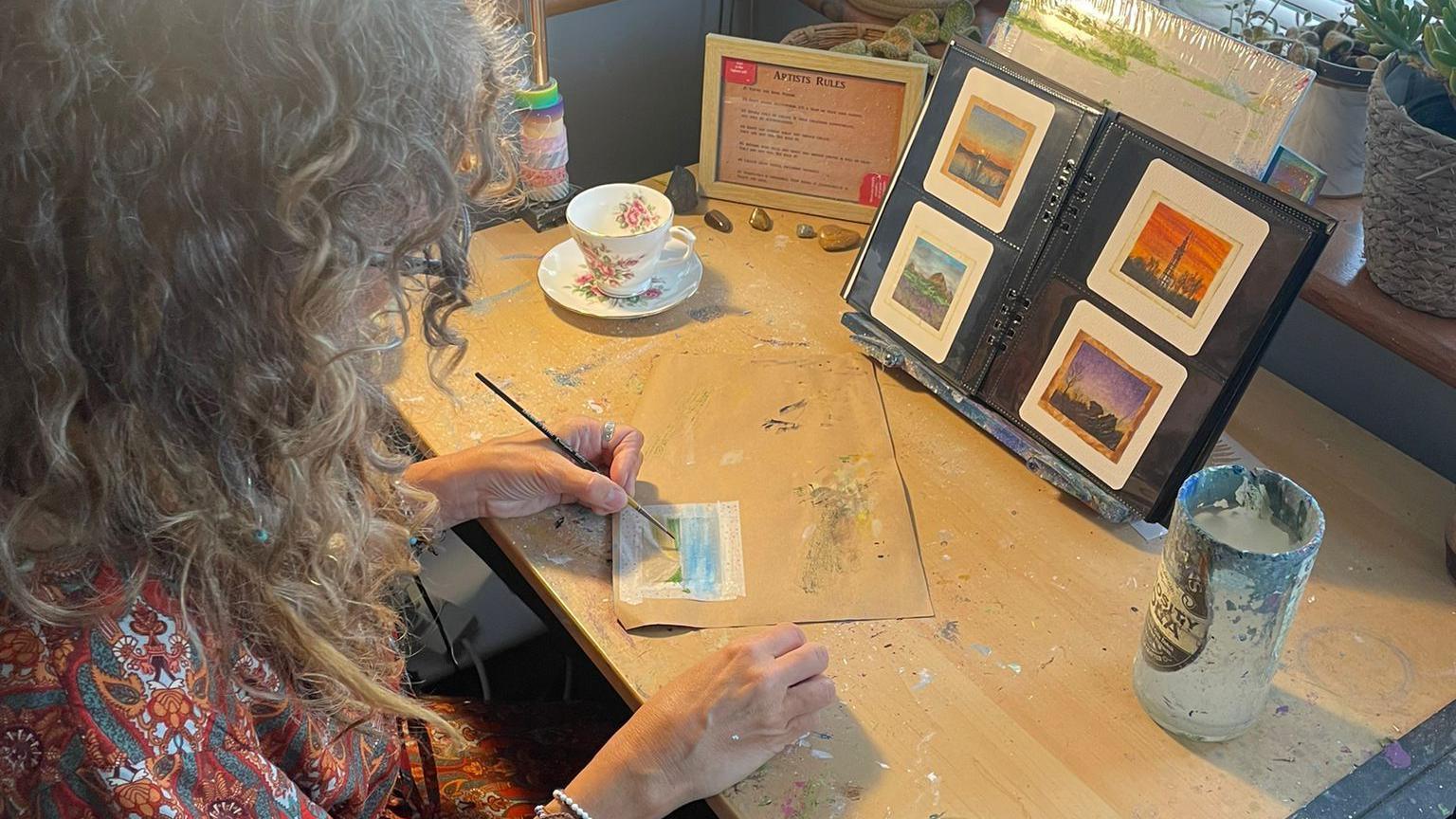 Ms West sitting at a desk holding a paintbrush. She is looking down and the camera is behind and above her so we can only see the back and side of her head. In front of her is a teabag with a painting on it. On the desk is a cup and saucer, a pot for rinsing her paintbrush and a binder with painted teabags displayed in it