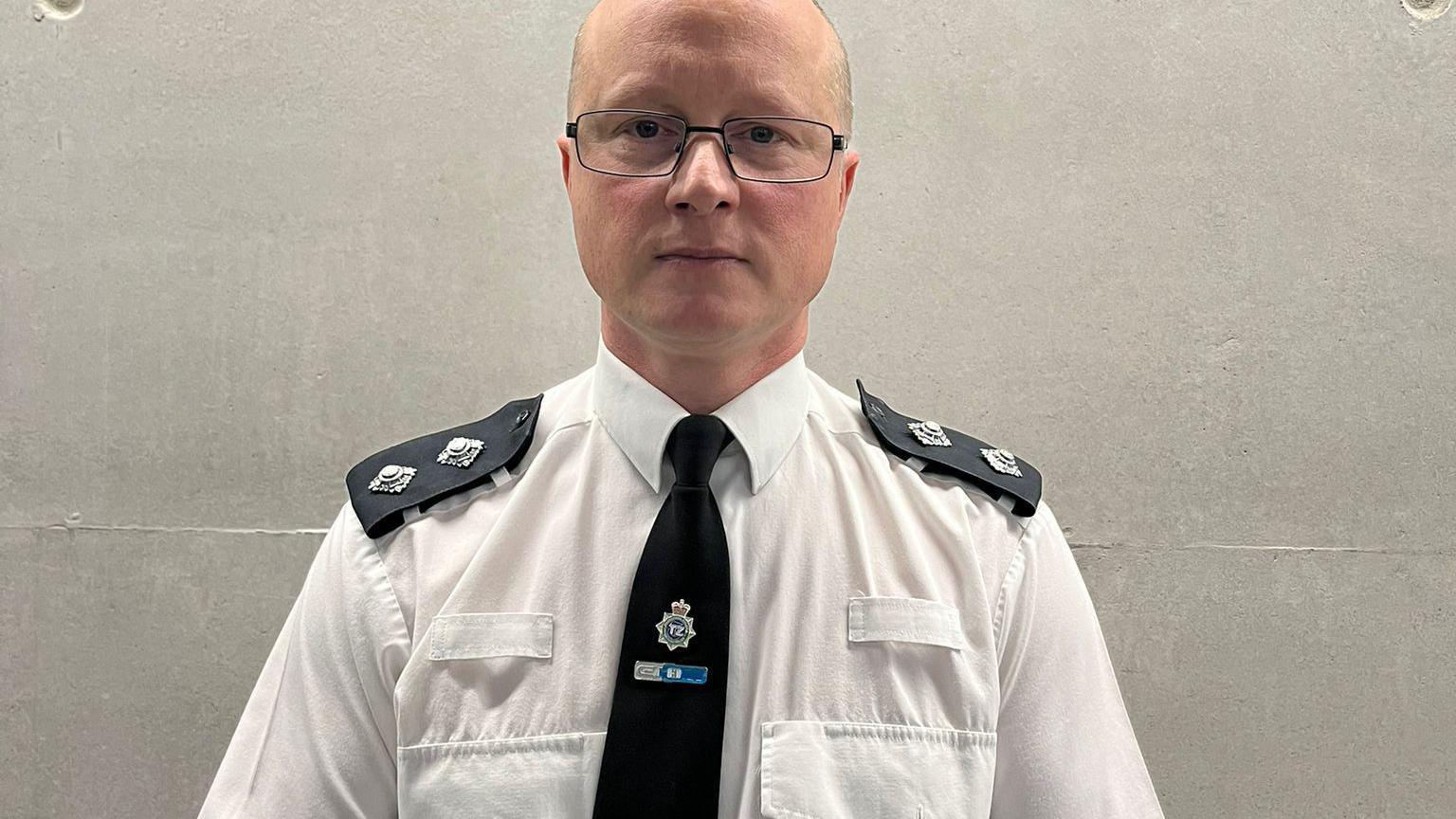 Insp Gavin Dixon, who has a bald head, glasses and wears a white police shirt with epaulettes and a black tie adorned with a Merseyside Police badge, looks into the camera with a serious expression