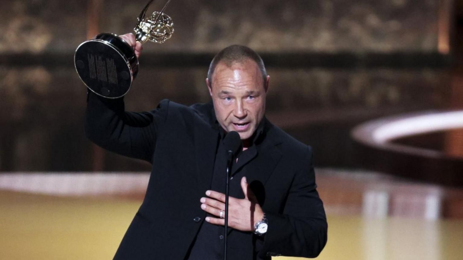Stephen Graham has short hair and is wearing a black suit and black shirt. He is holding an Emmy award aloft with his right hand and had his left hand on his chest.