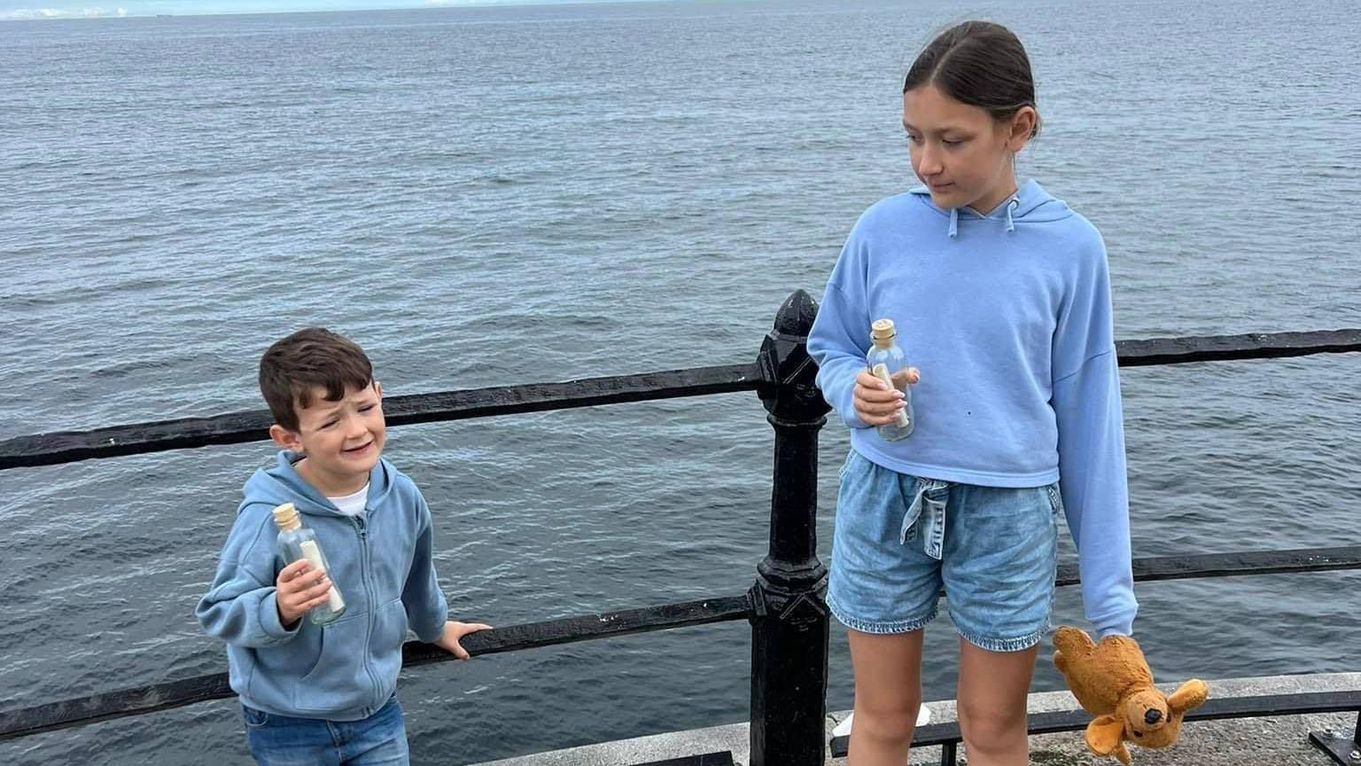 Harry and Grace holding their bottle at the pier, with the sea behind them. Harry is smiling at the camera and Grace is looking at him.