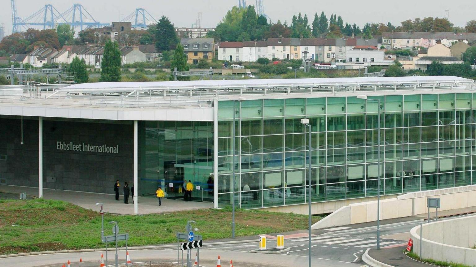 A train station with a series of glass window exterior, and a road in front of it. There is a sign on the left wall which reads "Ebbsfleet International" in large white letters.