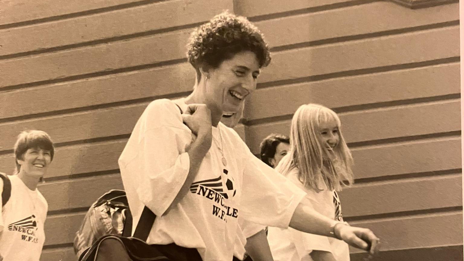 Margaret Hodgson, with short curly hair, smiles as she walks along a path, wearing a t-shirt that says Newcastle W.F.C, three other women walk beside her. 