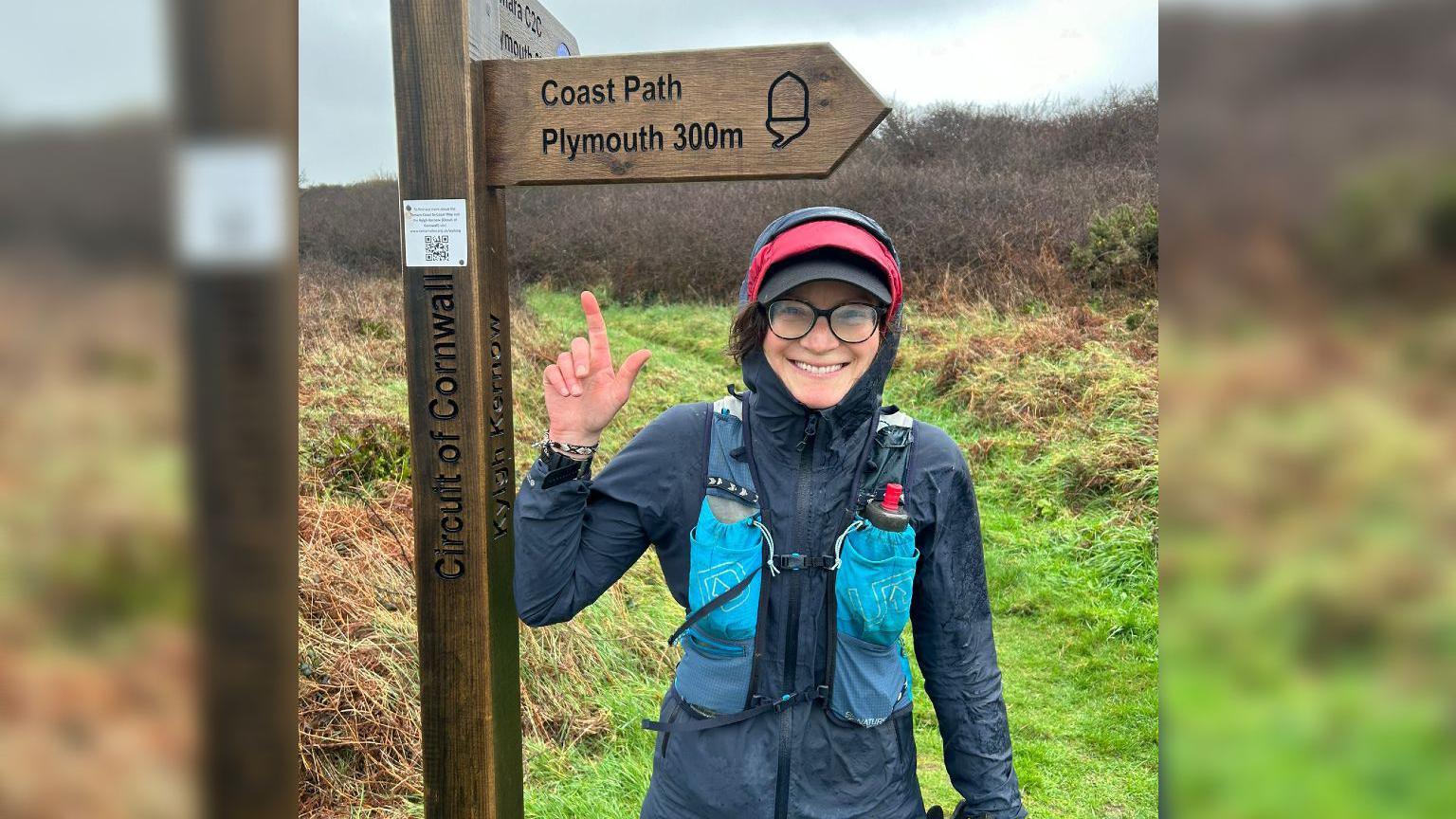 Claire Allen standing beside a Cornish coast path sign and pointing at it. It says Plymouth is 300m away. It is a wet and rainy day, she is wearing a raincoat, hat, and carrying a bottle of water in her runners backpack