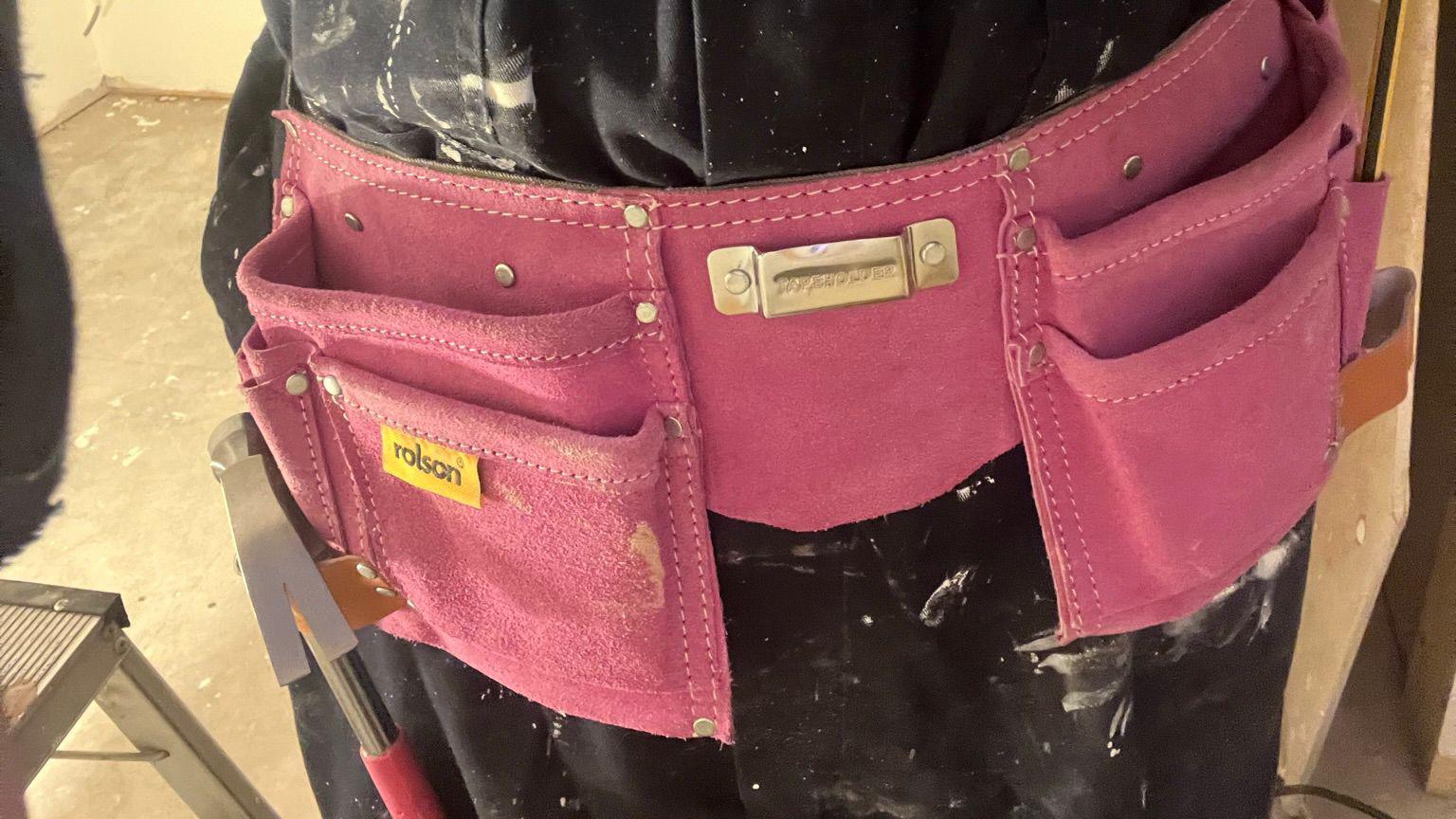 A pink suede toolbelt, with a hammer attached is on top of messy, white paint stained dark overalls 