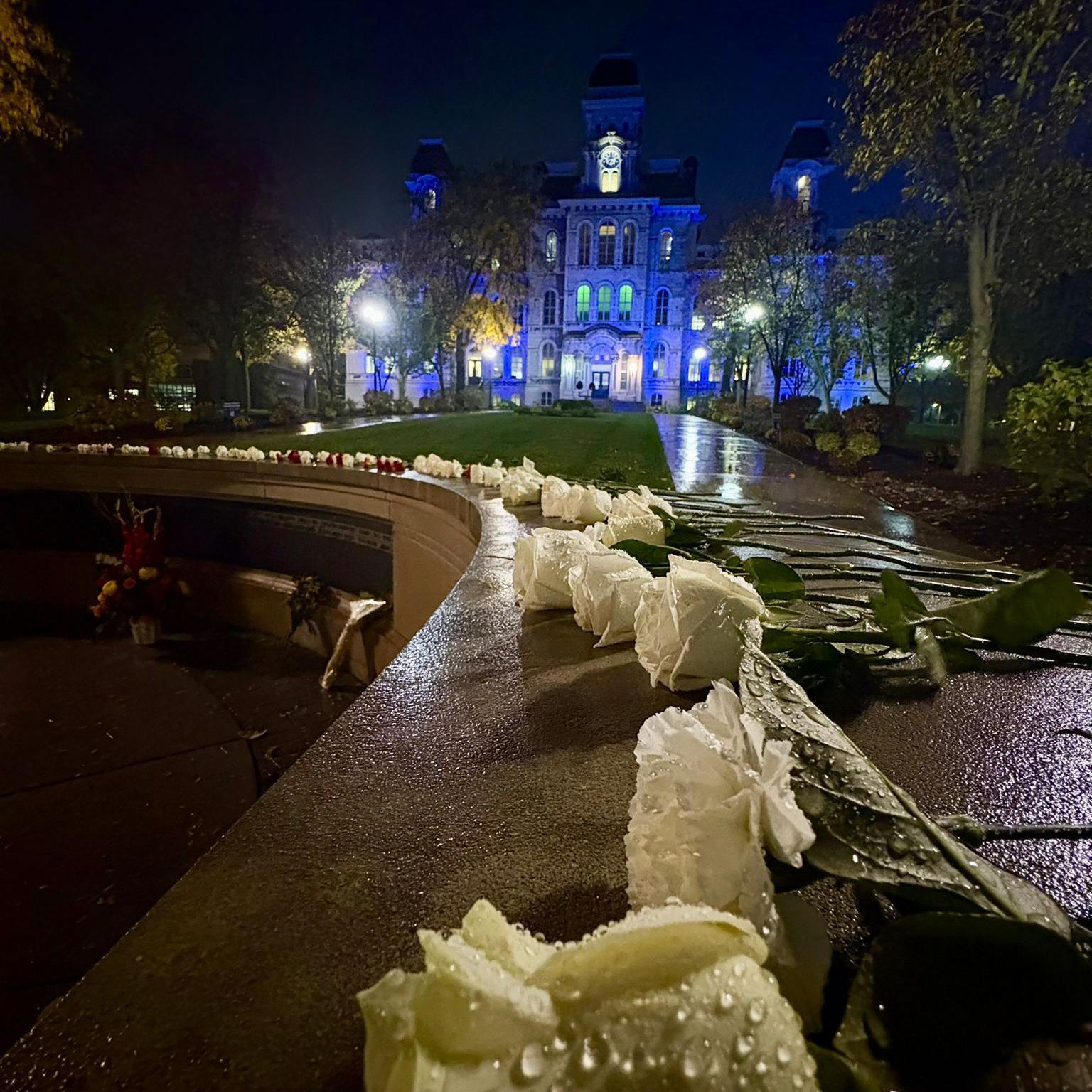 Dozens of white roses lie on top of a smooth wall, in front of a grand stone building. It is dark, the ground is wet and there are droplets of water visible on the flower petals.
