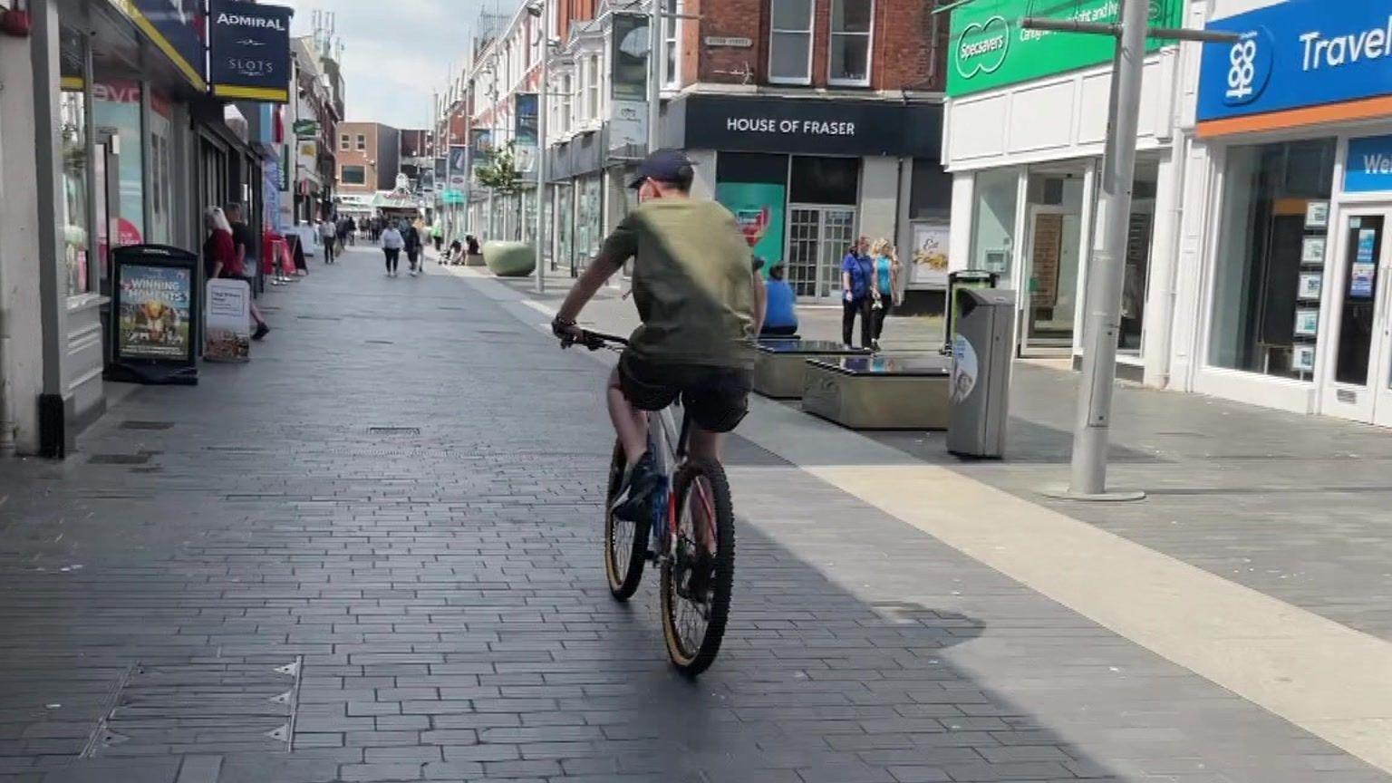 A man, wearing a green jumper and a black cap, cycles in a pedestrianised zone in Grimsby. Shops, including House of Fraser and Coop Travel, line the street on either side of him