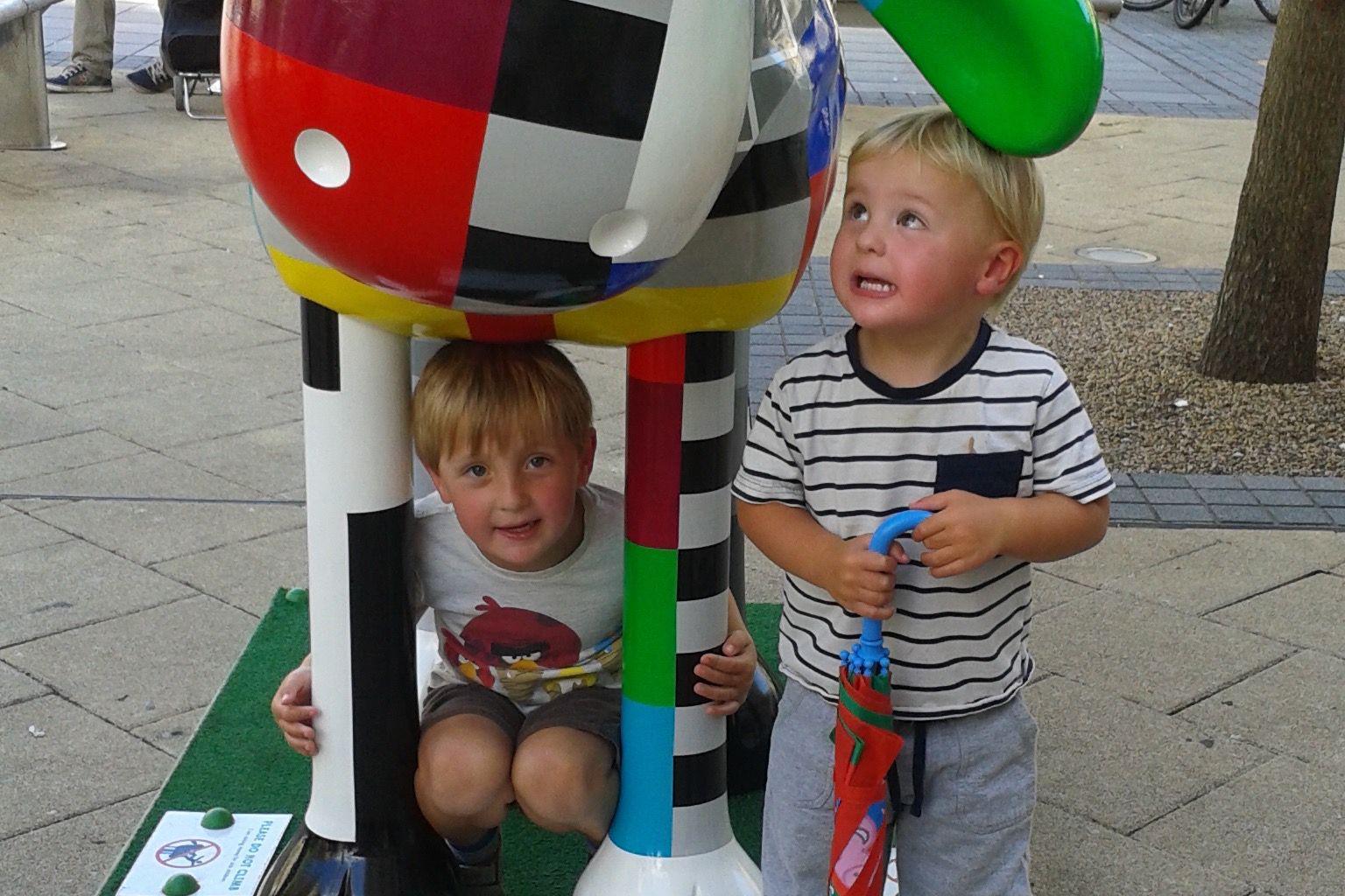 A young Leo and Hugh crouch beneath a large, colourful sculpture during the 2015 trail. The sculpture is covered in bright designs and patterns. The boys smile beneath its large, floppy ears.