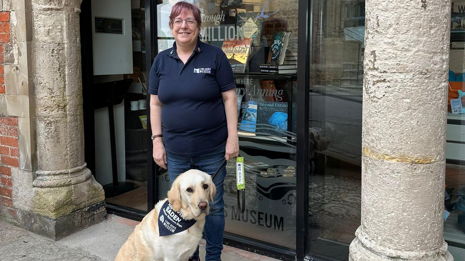 Lizzie Wiscombe with her guide dog posing for the camera between the stone pillars at the entrance of Lyme Regis Museum