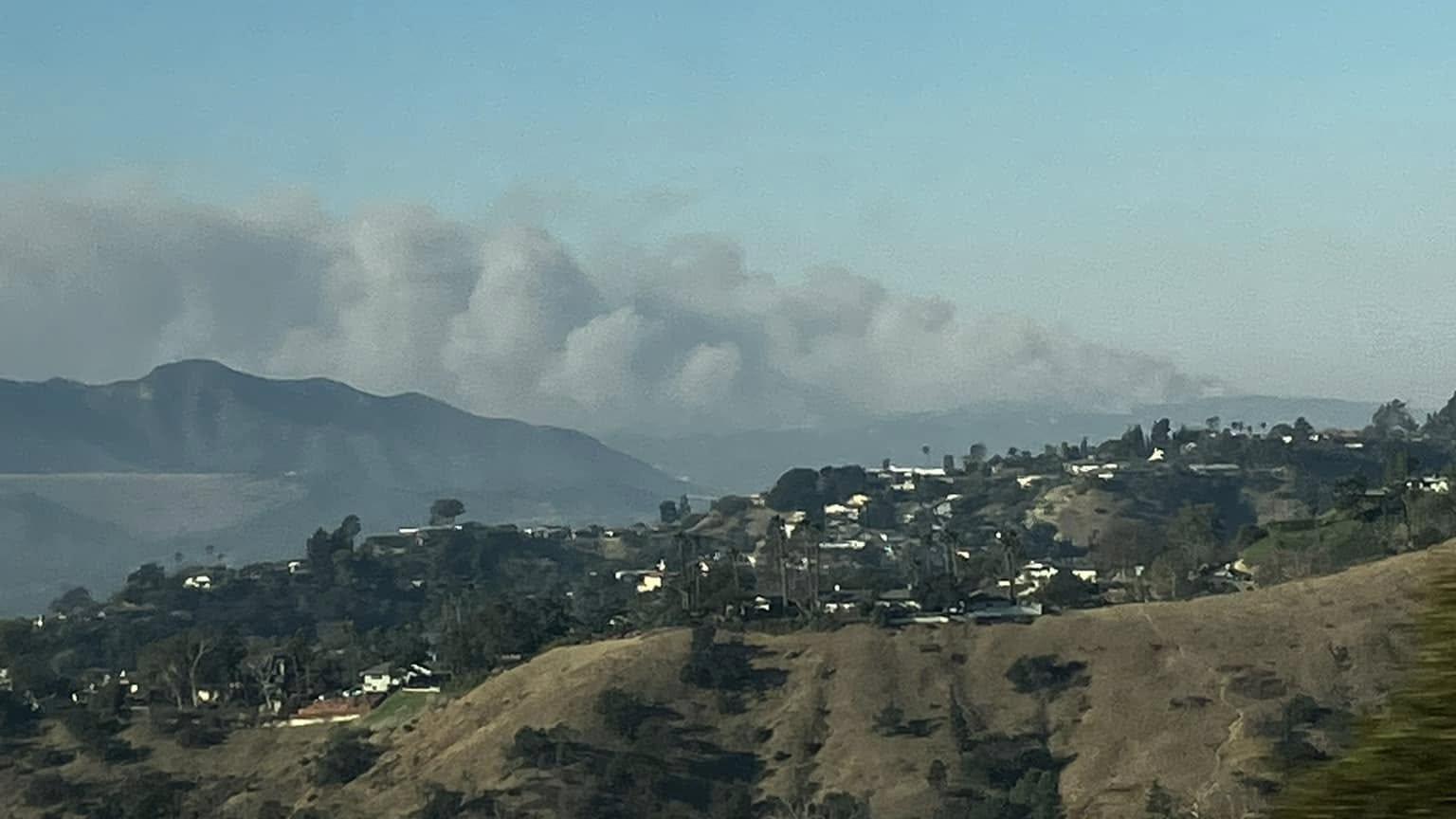 A clear blue sky, almost half covered by a large cloud of thick, dark grey smoke over Los Angeles